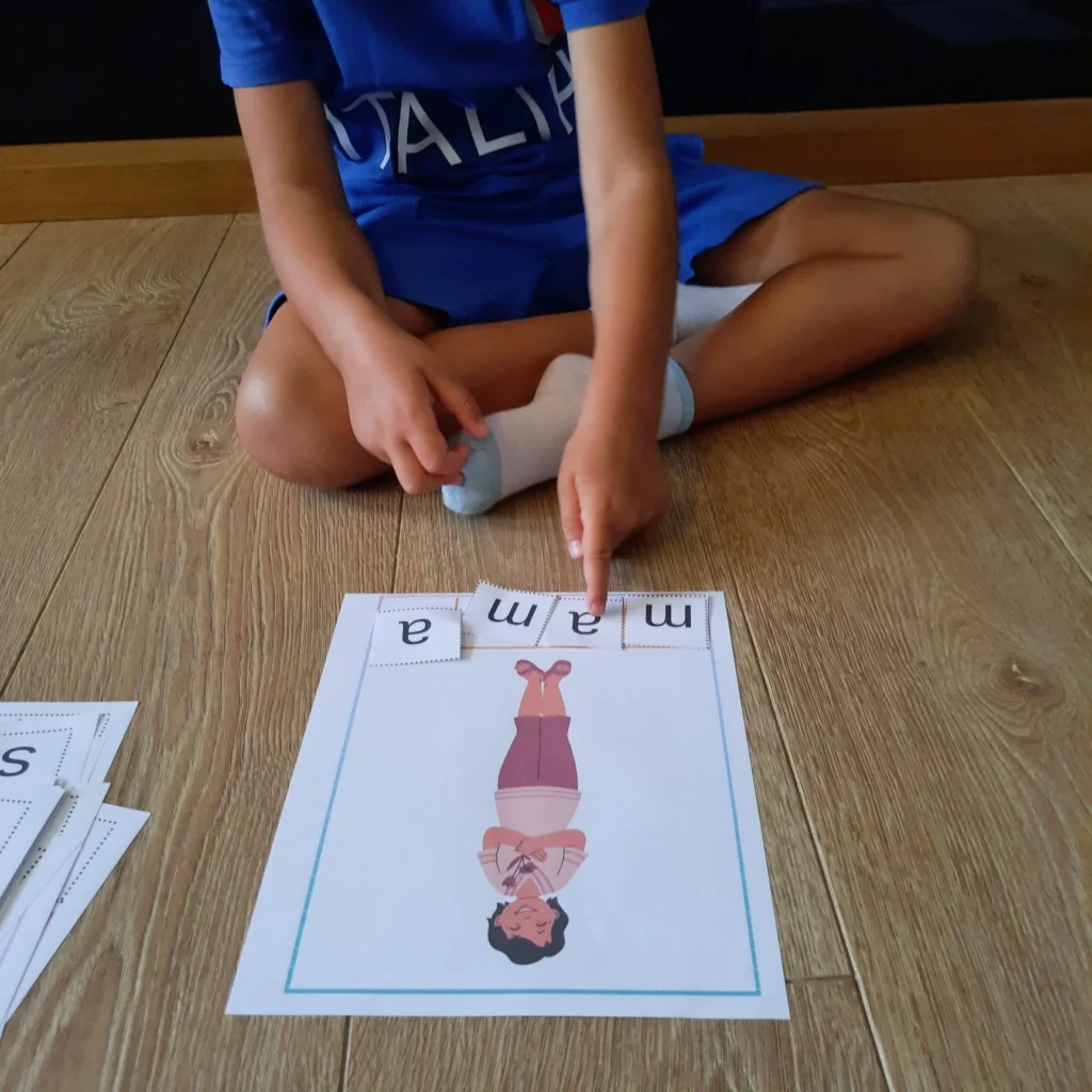 Dziecko siedzi na podłodze i układa litery tworzące wyraz „mama” przy ilustracji kobiety odwróconej do góry nogami.