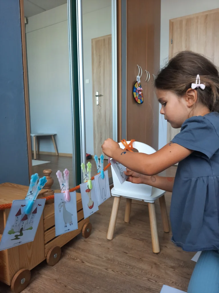 Dziewczynka przypina kolorowymi klamerkami karty edukacyjne z rysunkami do pomarańczowego sznurka rozciągniętego w pokoju.