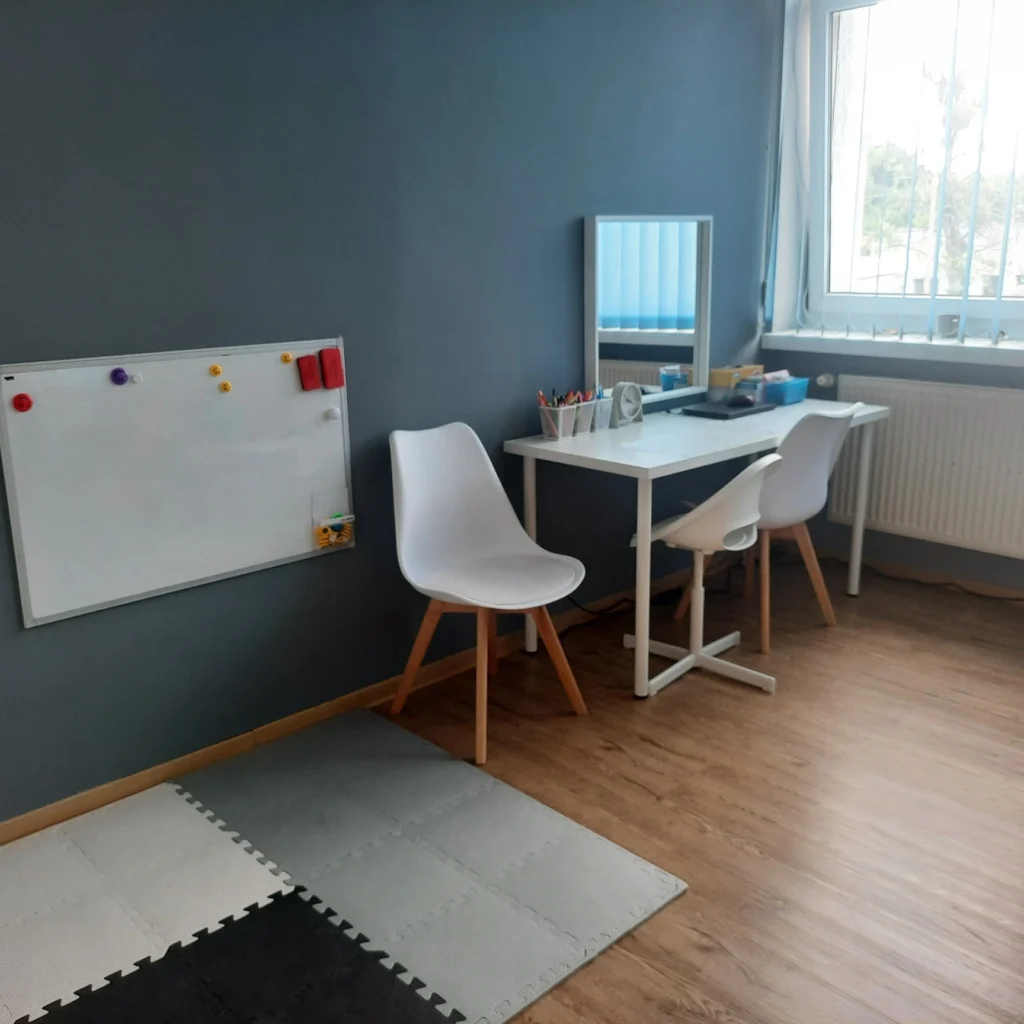 Gabinet terapeutyczny z biurkiem, tablicą suchościeralną i matą do ćwiczeń przygotowany do zajęć z dziećmi.