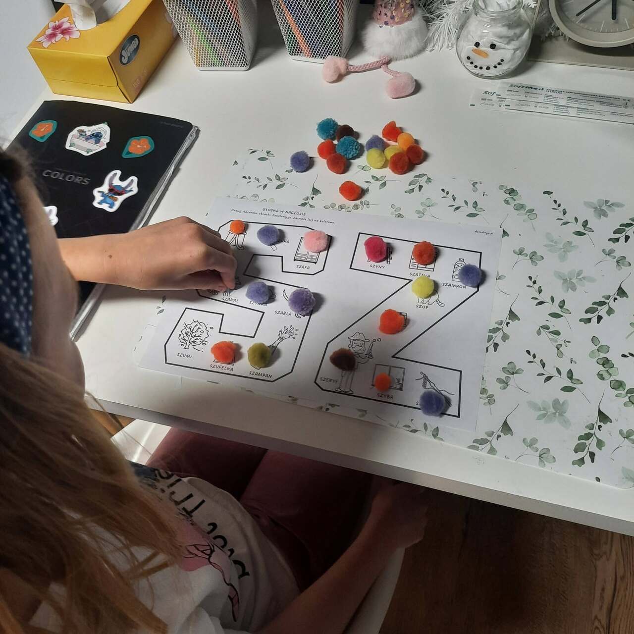 Dziecko podczas terapii logopedycznej wykonuje ćwiczenie rozwijające słownictwo i głoskę „SZ”, dopasowując kolorowe pompony do ilustracji na karcie pracy.